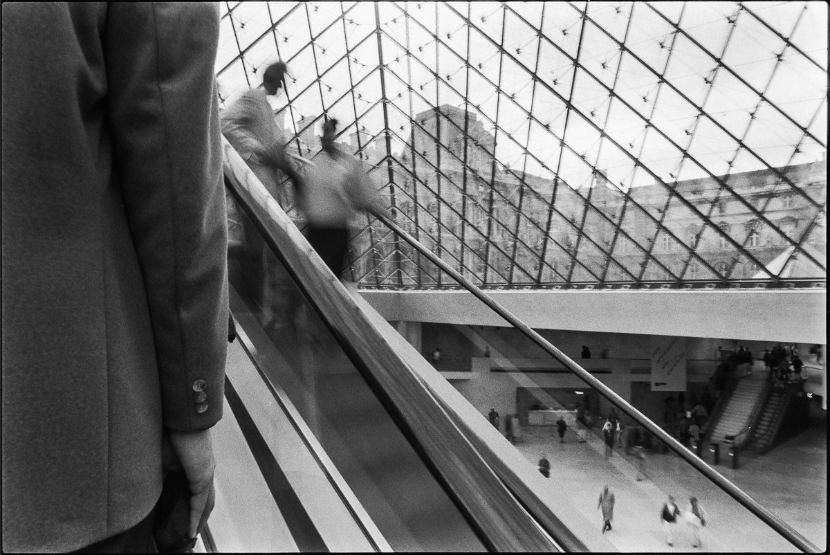 Escalator du Louvre sous la pyramide en 1989, silhouettes floues et diagonales architecturales en noir et blanc